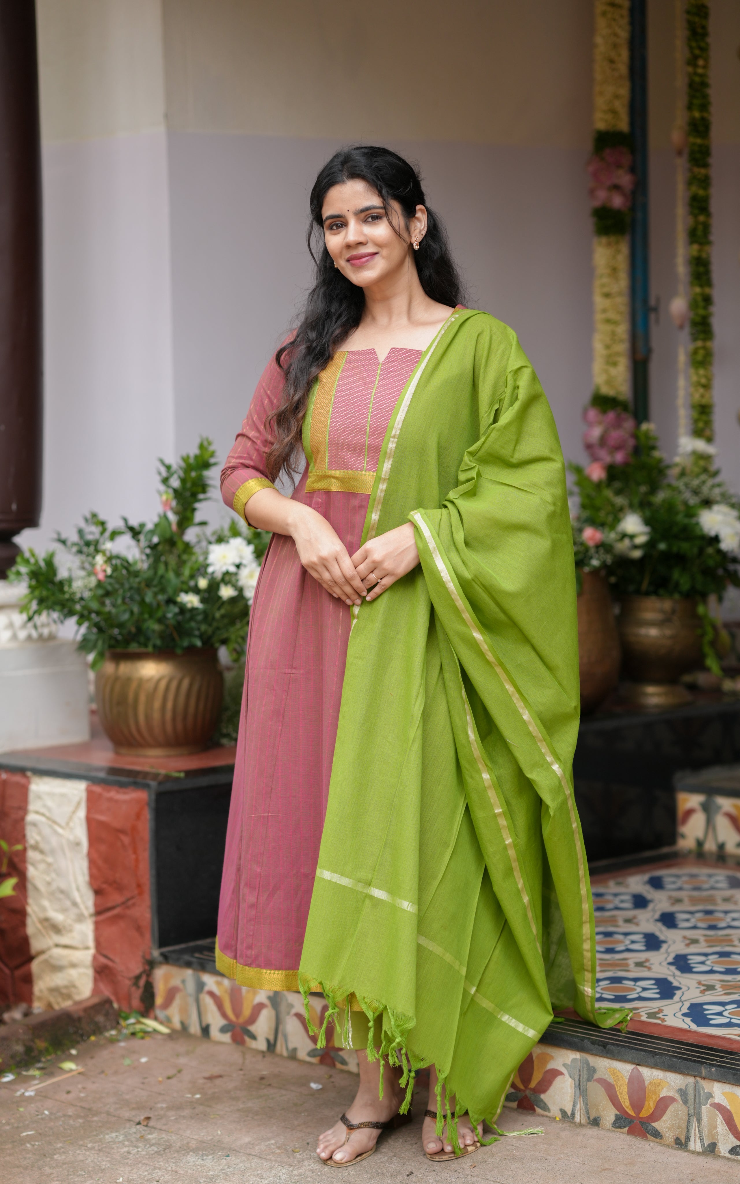 A woman standing outdoors wearing a pink flared South Cotton Anarkali paired with a contrasting green dupatta